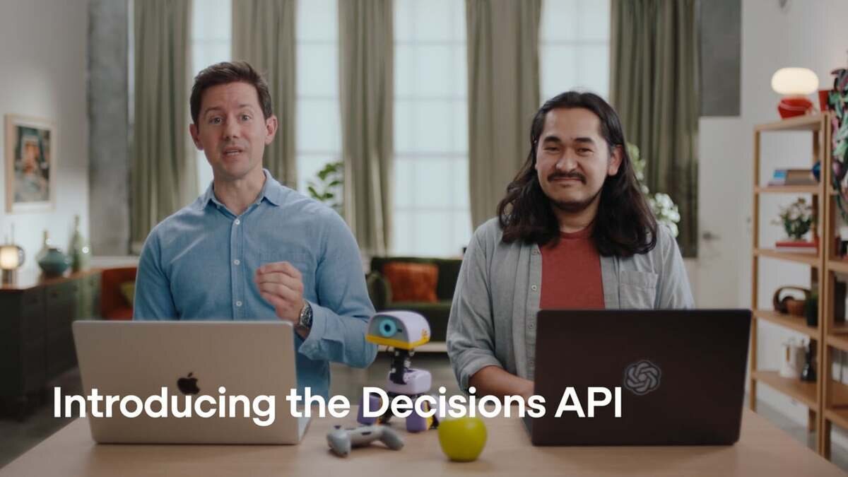 Pôster do vídeo de lançamento da Decisions API: dois homens sentados à mesa com notebooks, um deles com o logo da OpenAI, e o texto Introducing the Decisions API em branco na base.