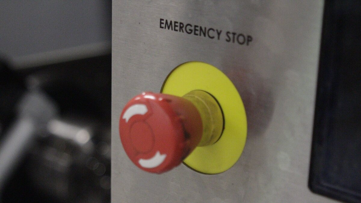 Fotografia em close de um botão de emergência industrial: o cogumelo vermelho com duas setas brancas curvas sobre anel amarelo, montado em painel de aço escovado, e o rótulo EMERGENCY STOP em preto acima do botão.